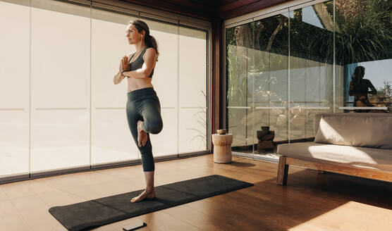 Mature woman practicing the tree pose during a yoga session. Senior woman doing a balancing asana on an exercise mat. Active woman following a healthy workout routine at home. - JLPSF09836