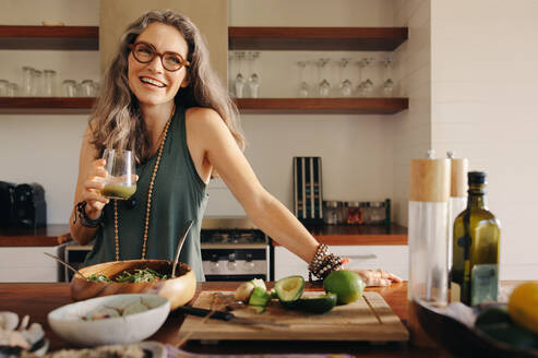 Healthy senior woman smiling while holding some green juice in her kitchen. Mature woman serving herself wholesome vegan food at home. Happy woman taking care of her aging body with a plant-based diet. - JLPSF09850