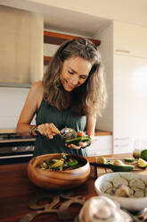 Glückliche Vegetarierin, die lächelnd Avocado in ihre Buddha-Schüssel gibt. Ältere Frau, die sich zu Hause eine köstliche vegane Mahlzeit serviert. Frau, die ihren alternden Körper mit einer gesunden pflanzlichen Ernährung pflegt. - JLPSF09860