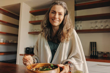 Mature woman smiling at the camera while having a healthy vegan meal. Happy senior woman serving herself a wholesome buddha bowl at home. Woman taking care of her aging body with a plant-based diet. - JLPSF09863