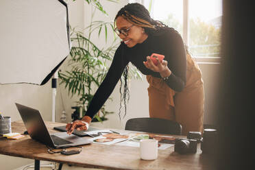 Freiberuflerin, die einen Anruf in ihrem Büro zu Hause entgegennimmt. Glückliche Fotografin, die fröhlich lächelt, während sie einen Laptop benutzt. Kreative junge Frau, die mit ihren Kunden kommuniziert, während sie an einem neuen Projekt arbeitet. - JLPSF09867