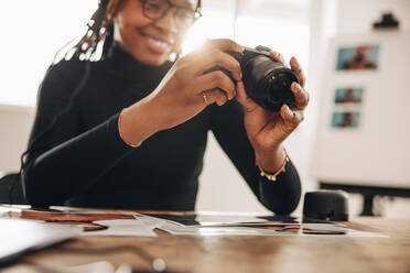 Fotografin hält eine DSLR-Kamera in ihrer Kamera in ihrem Büro zu Hause. Kreative junge Frau lächelt fröhlich, während sie an ihrem Schreibtisch arbeitet. Freiberuflerin arbeitet an einem neuen fotografischen Projekt. - JLPSF09872