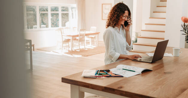 Geschäftsfrau zu Hause arbeitet am Laptop und spricht über Handy. Weibliche Unternehmerin sitzt am Tisch und telefoniert, während sie auf einen Laptop-Computer schaut. - JLPSF09878