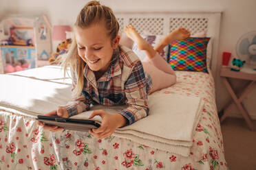Junges Mädchen liegt auf dem Bett und benutzt ein digitales Tablet. Junges Mädchen entspannt sich in ihrem Zimmer und schreibt einer Freundin eine SMS auf ihrem Tablet-PC. - JLPSF09883