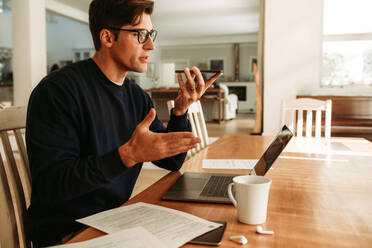 Businessman working from at home talking on speaker phone. Man sitting at table with a laptop making a phone call. - JLPSF09887