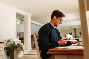 Lächelnder Mann am Tisch sitzend, der mit einem Handy in der Hand am Laptop arbeitet. Glücklicher Geschäftsmann, der von zu Hause aus arbeitet. - JLPSF09888