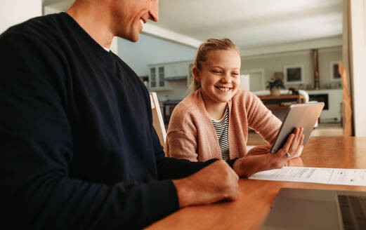 Vater und Tochter sitzen am Tisch und benutzen gemeinsam ein digitales Tablet zu Hause. Mädchen schaut auf den Tablet-Computer in der Hand ihres Vaters zu Hause. - JLPSF09890