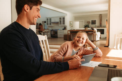 Mann mit seiner Tochter am Tisch sitzend und lächelnd. Mann hält ein digitales Tablet und spricht mit seiner Tochter zu Hause. - JLPSF09893
