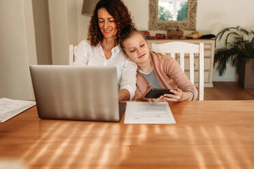 Girl using mobile phone while sitting close to her mother working on a laptop. Woman working from home sitting with her daughter using a smart phone. - JLPSF09894