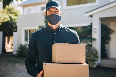 Delivery man with face mask delivering parcel boxes. Male courier worker wearing face mask and cap holding cardboard boxes outdoors. - JLPSF09900