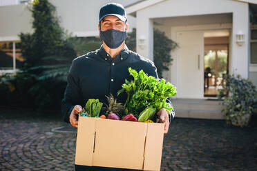 Zusteller mit Gesichtsmaske und Lebensmittelkarton im Freien, der eine Kiste mit Obst und Gemüse an eine Privatadresse ausliefert. - JLPSF09902