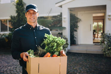 Man holding a box of fruits and vegetables outdoors looking at camera. Delivery person delivering online ordered grocery to home address. - JLPSF09903