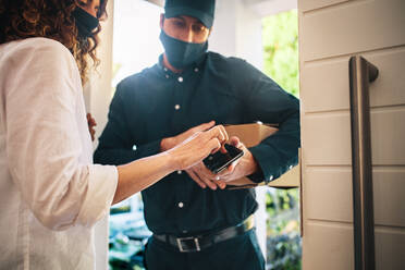 Ein Zusteller mit Gesichtsmaske hält ein Smartphone in der Hand, während eine Kundin auf dem Touchscreen die Quittung für ein Zustellungspaket unterschreibt. - JLPSF09910