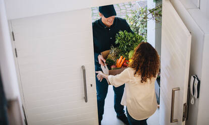 High angle view of a female customer receiving groceries from delivery man at the house's door. Delivery employee delivering a box of fruits and vegetables to a woman. - JLPSF09912