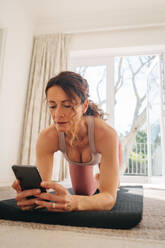 Frau, die ihr Smartphone benutzt, um online Trainingsanleitungen zu finden, während sie zu Hause trainiert. Frau, die auf ihrem Telefon online Yoga-Kurse schaut, während sie trainiert. - JLPSF09932