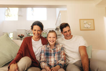 Happy family of three sitting on a sofa and watching TV together at home. Girl with her father and mother sitting on couch watching television and laughing. - JLPSF09950