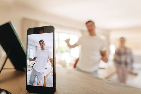 Man dancing in front of a mobile phone with family at the back. Man creating his dancing video on a mobile app to share on his social media. - JLPSF09955