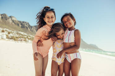 Drei bezaubernde kleine Mädchen, die sich lächelnd umarmen, während sie am Strand stehen. Eine Gruppe von glücklichen jungen Freunden, die während der Sommerferien zusammen Spaß haben. - JLPSF09956