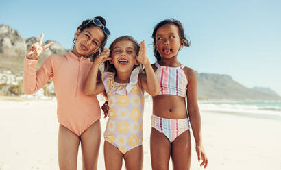 Verspielte kleine Mädchen, die lustige Gesichter machen, während sie zusammen am Strand stehen. Gruppe von entzückenden jungen Freunden, die während der Sommerferien zusammen Spaß haben. - JLPSF09957