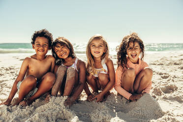 Niedliche kleine Kinder, die in die Kamera lächeln, während sie am Strand im Sand sitzen. Eine Gruppe fröhlicher junger Kinder, die sich in den Sommerferien amüsieren. - JLPSF09961