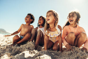 Sommerspaß am Strand. Vier junge Freunde lachen fröhlich, während sie am Strand auf dem Sand sitzen. Eine Gruppe liebenswerter kleiner Kinder, die in den Sommerferien eine gute Zeit miteinander verbringen. - JLPSF09962