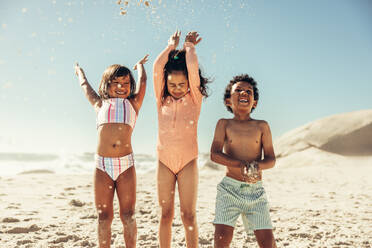 Fröhliche Kinder haben Spaß, während sie Strandsand in die Luft werfen. Eine Gruppe von drei bezaubernden kleinen Kindern, die ihren Sommerurlaub an einem sonnigen Strand genießen. - JLPSF09966