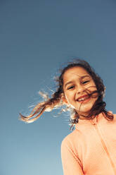 Glückliches kleines Mädchen, das in die Kamera lächelt, während es mit dem Himmel im Hintergrund steht. Liebenswertes junges Mädchen, das im Sommer am Strand Spaß hat. - JLPSF09984