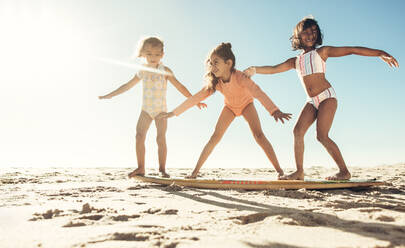 Drei glückliche kleine Mädchen, die am Strand auf einem Surfbrett spielen. Eine Gruppe liebenswerter kleiner Kinder, die in den Sommerferien zusammen Spaß haben. - JLPSF09987