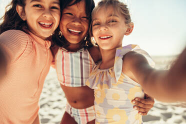 Drei glückliche kleine Mädchen, die für ein Gruppen-Selfie am Meer lächeln. Eine Gruppe liebenswerter kleiner Kinder, die in den Sommerferien zusammen Spaß haben. - JLPSF09991