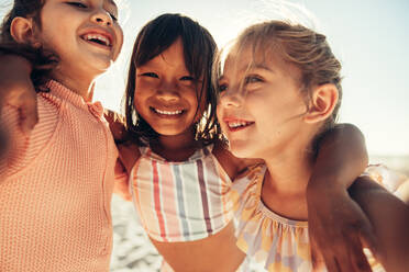 Drei glückliche kleine Mädchen, die für ein Gruppen-Selfie am Strand lächeln. Eine Gruppe liebenswerter kleiner Kinder, die in den Sommerferien zusammen Spaß haben. - JLPSF09992