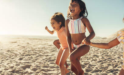 Aufgeregte kleine Mädchen, die zusammen am Strand rennen und Spaß haben. Eine Gruppe glücklicher kleiner Kinder, die ihren Sommerurlaub an einem sonnigen Strand genießen. - JLPSF09996