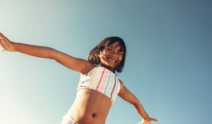 Flying high in the sky. Low angle view of an adorable little girl smiling at the camera with her arms outstretched. Happy young girl having fun at the beach during summer vacation. - JLPSF09999