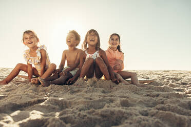 Nette kleine Kinder, die fröhlich lachen, während sie zusammen am Strand sitzen. Eine Gruppe unbeschwerter junger Freunde, die in den Sommerferien zusammen Spaß haben. - JLPSF10001
