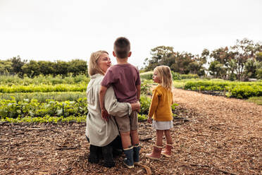 Junge alleinerziehende Mutter im Gespräch mit ihren Kindern auf einem Biobauernhof. Zweifache Mutter kniet mit ihren Kindern vor frischem Grün. Rückansicht einer jungen, sich selbst versorgenden Familie auf einem landwirtschaftlichen Feld. - JLPSF10010