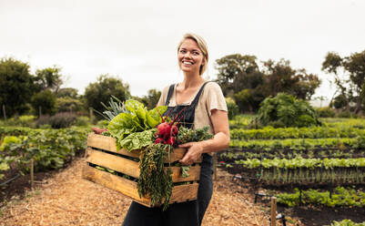 Glückliche Bäuerin, die eine Kiste mit frischem Obst und Gemüse in der Hand hält. Junge Bio-Bäuerin, die nach der Ernte von frischem Gemüse aus ihrem Garten fröhlich lächelt. Erfolgreiche Bäuerin, die auf ihrem Hof steht. - JLPSF10050