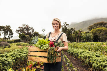 Lächelnde Bäuerin, die eine Kiste mit frischem Obst und Gemüse in der Hand hält. Junge Biobäuerin, die in die Kamera schaut, nachdem sie frisches Gemüse aus ihrem Garten geerntet hat. Glückliche Bäuerin, die auf ihrem Hof steht. - JLPSF10053