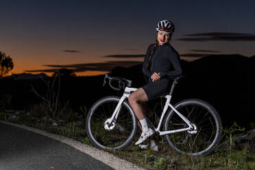 Lächelnder Radfahrer, der sich in der Abenddämmerung auf ein Fahrrad in der Nähe der Straße stützt - AMNF00020