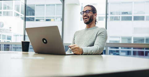 Glücklicher junger Geschäftsmann lacht fröhlich, während er mit einem Laptop arbeitet. Erfolgreicher Unternehmer, der aus der Ferne in einem Co-Working Space arbeitet. - JLPSF10119