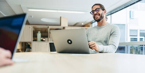 Glücklicher junger Geschäftsmann, der fröhlich lächelt, während er mit einem Laptop arbeitet. Erfolgreicher Unternehmer, der aus der Ferne in einem Co-Working Space arbeitet. - JLPSF10121