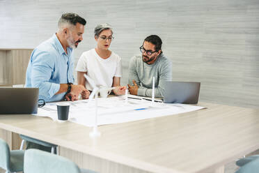 Drei kreative Konstrukteure, die in einem modernen Büro mit Plänen für Windkraftanlagen arbeiten. Ein Team innovativer Geschäftsleute beim Brainstorming, während sie an einem Projekt für erneuerbare Energien arbeiten. - JLPSF10128