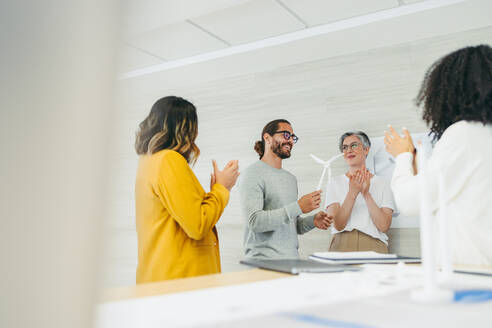 Successful designers applauding their colleague during a meeting in a modern office. Group of innovative businesspeople celebrating their teammate's achievement in a wind turbine project. - JLPSF10137