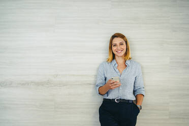 Happy young businesswoman smiling cheerfully while holding a smartphone in a modern office. Female entrepreneur sending a text message to her business associates. - JLPSF10159