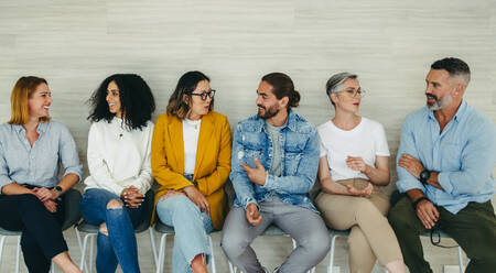 Cheerful businesspeople talking to each other while sitting in line for an interview. Group of successful job applicants sharing ideas in a modern workplace. - JLPSF10177