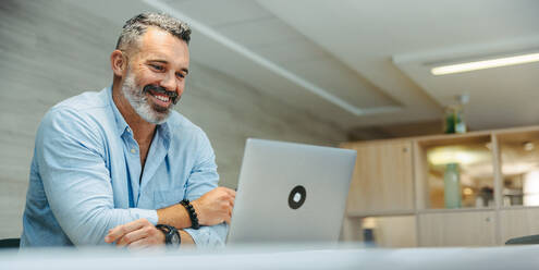 Cheerful entrepreneur attending an online meeting with his business associates. Happy mature businessman having a video conference while working remotely in a modern co-working office. - JLPSF10189