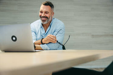 Happy mature businessman attending an online meeting with his business associates. Cheerful entrepreneur having a video conference while working remotely in a modern co-working office. - JLPSF10190
