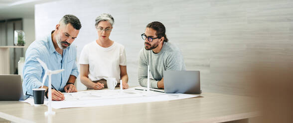 Ein Team von kreativen Geschäftsleuten fertigt Zeichnungen für Windkraftanlagen an. Innovative Design-Profis arbeiten in einem modernen Büro an einem Projekt für erneuerbare Energien und machen Brainstorming. - JLPSF10199