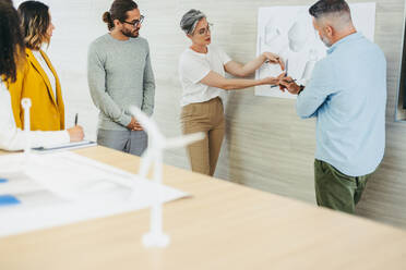 Mature design professional explaining some drawings during a meeting with her colleagues. Innovative businesspeople sharing creative ideas while working on a wind turbine project in a modern office. - JLPSF10201