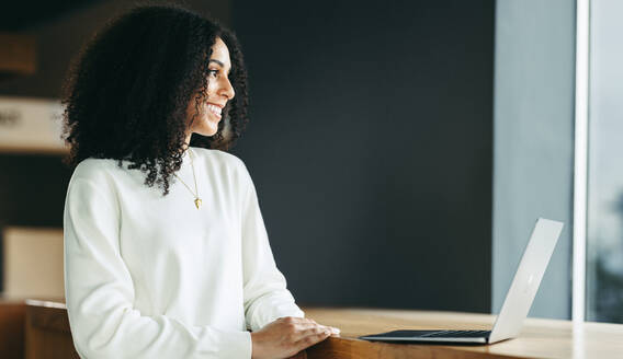 Glückliche junge Geschäftsfrau, die lächelt und wegschaut, während sie mit einem Laptop arbeitet. Fröhliche ethnische Geschäftsfrau, die aus der Ferne in einem modernen Co-Working Space arbeitet. - JLPSF10212