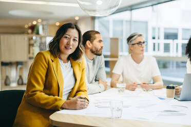 Fröhliche Geschäftsfrau, die in die Kamera schaut, während sie in einer Besprechung mit ihren Kollegen sitzt. Gruppe innovativer Design-Profis, die sich über kreative Ideen austauschen, während sie an einem neuen Projekt arbeiten. - JLPSF10223