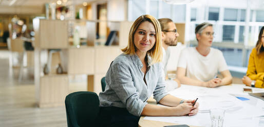 Selbstbewusste junge Designerin, die in die Kamera schaut, während sie in einer Besprechung mit ihrem Team sitzt. Eine Gruppe innovativer Geschäftsleute tauscht kreative Ideen aus, während sie an einem neuen Projekt arbeiten. - JLPSF10230